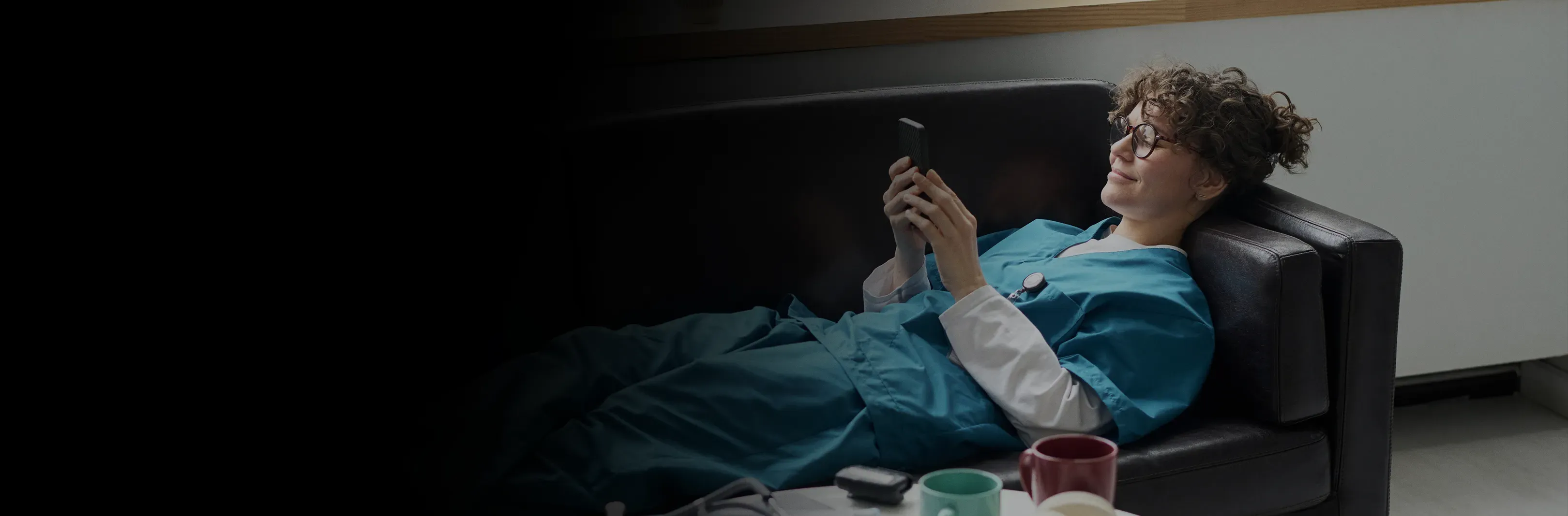 A woman in teal scrubs relaxes on a black leather sofa, smiling at her phone.
